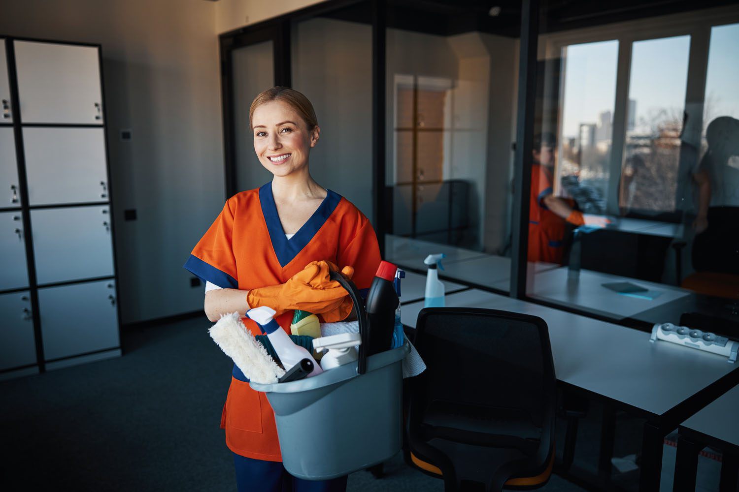 Reinigungskraft mit orangenem Shirt und Putzeimer lächelt in die Kamera.